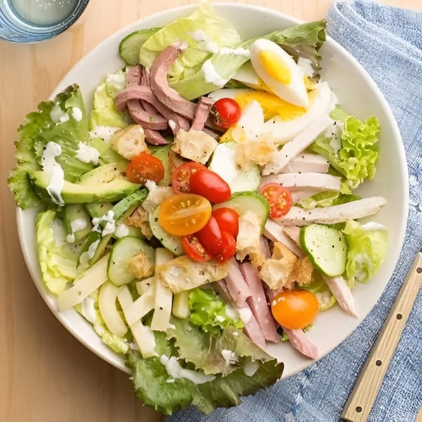 A vibrant deli salad with lettuce, meats, hard-boiled egg, tomatoes, cucumbers, avocado, and croutons.