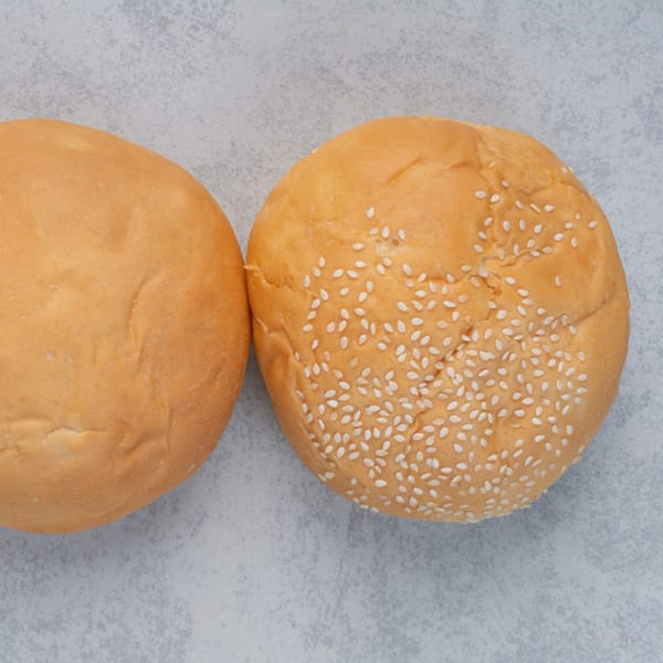 Two golden-brown burger buns, one plain and one with sesame seeds, on a light gray surface.